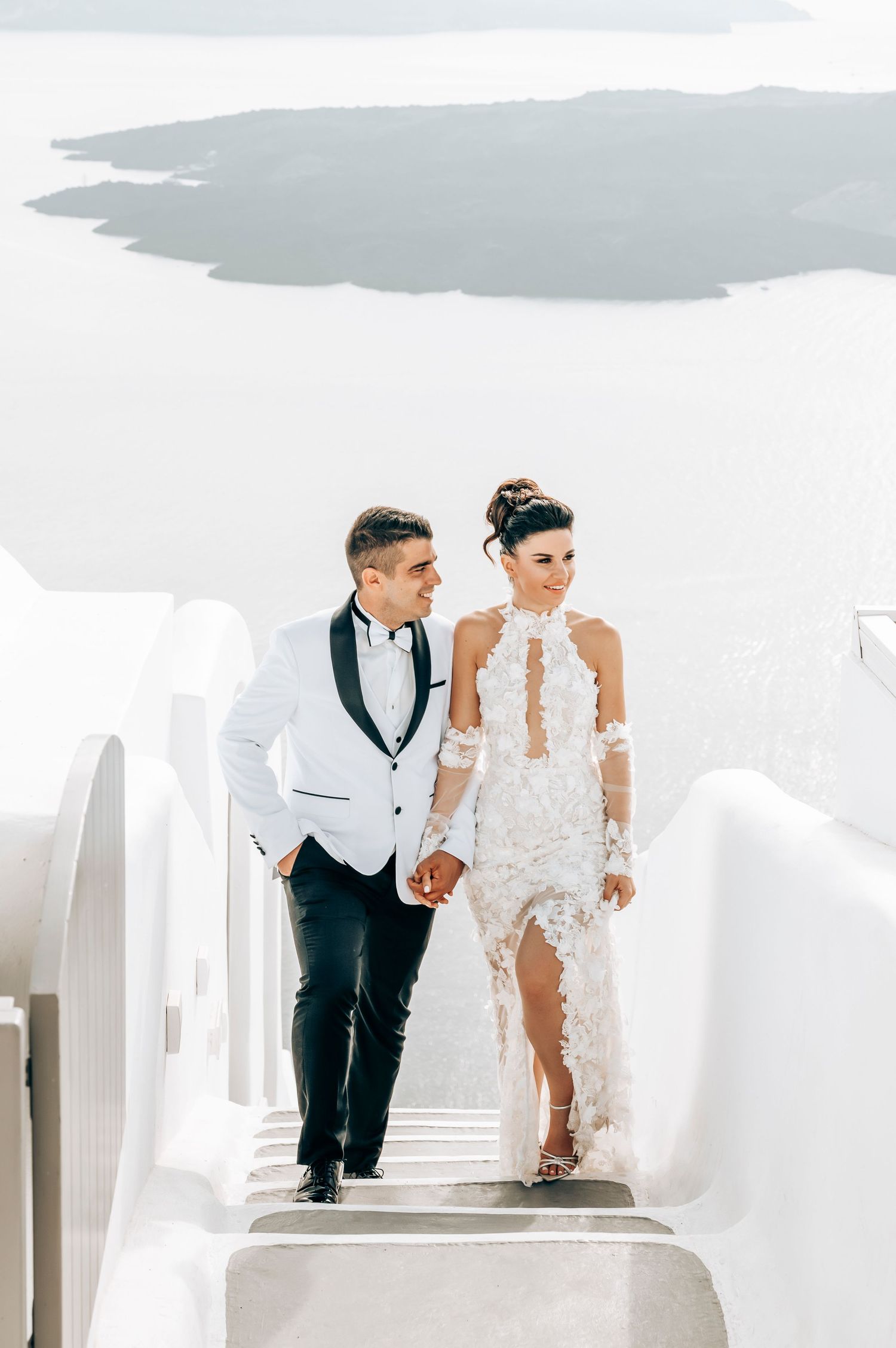 Newlyweds walking down white steps in Santorini with Mediterranean backdrop.