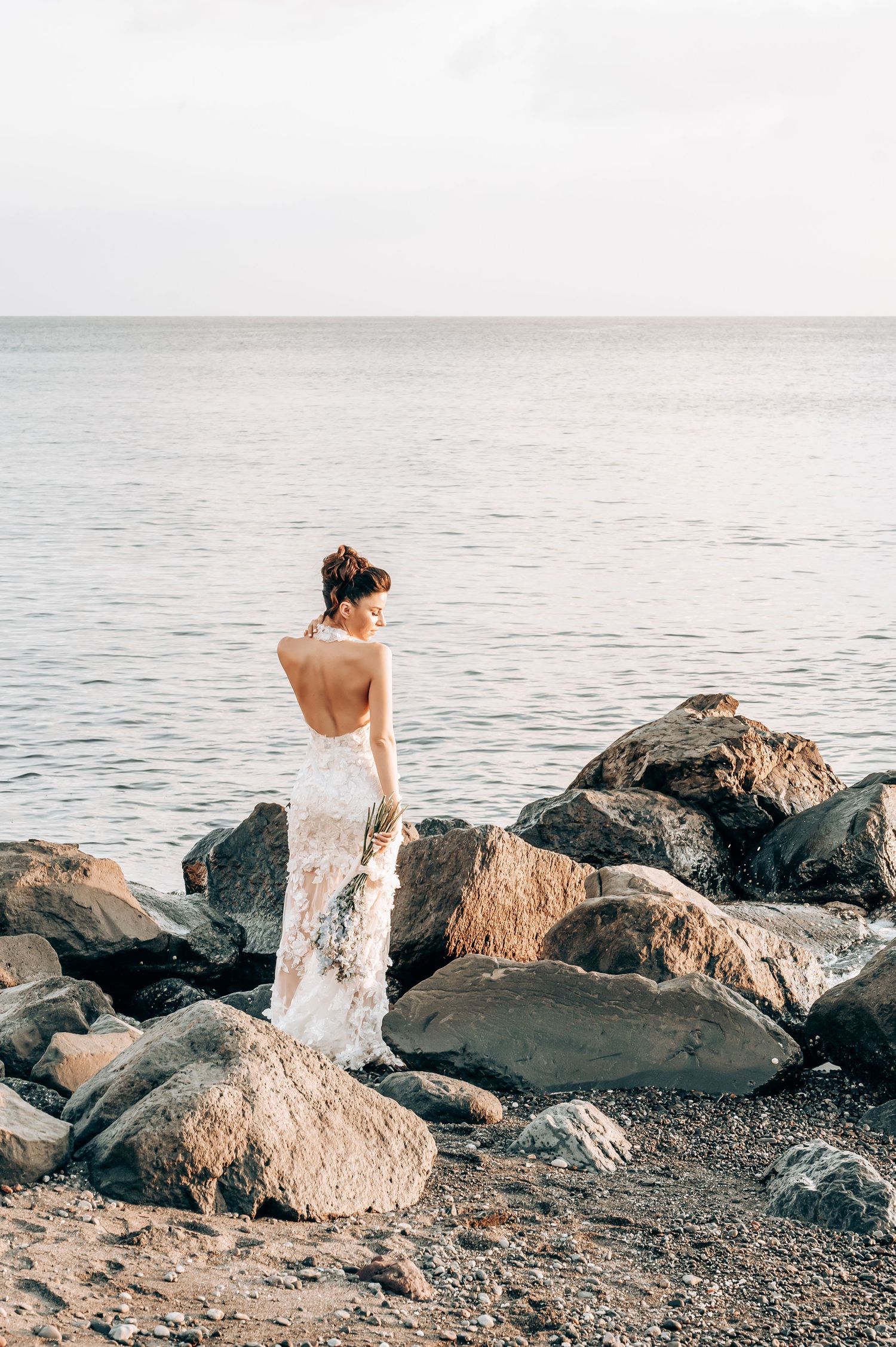 Bride in backless lace dress poses on rocky beach overlook.