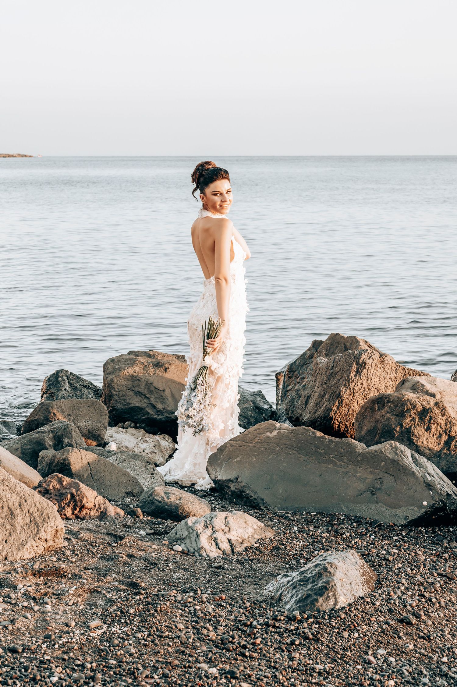 A bride in a lace gown poses on rocky coastal terrain against a serene ocean backdrop.