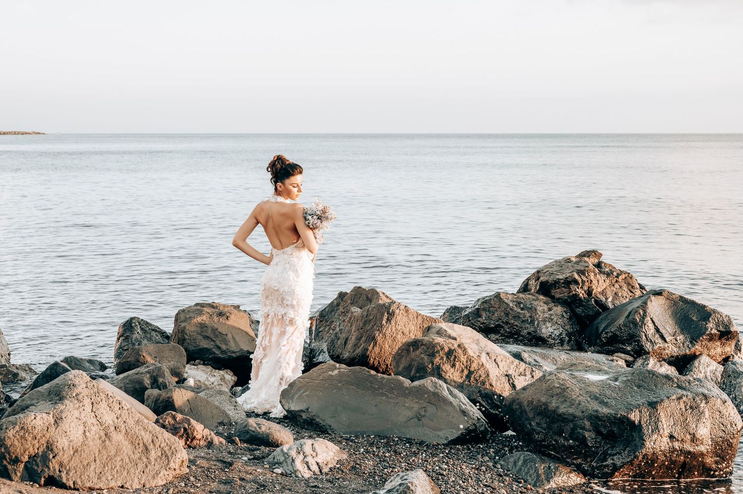 A backlit bride walks across coastal rocks in a delicate lace wedding dress.