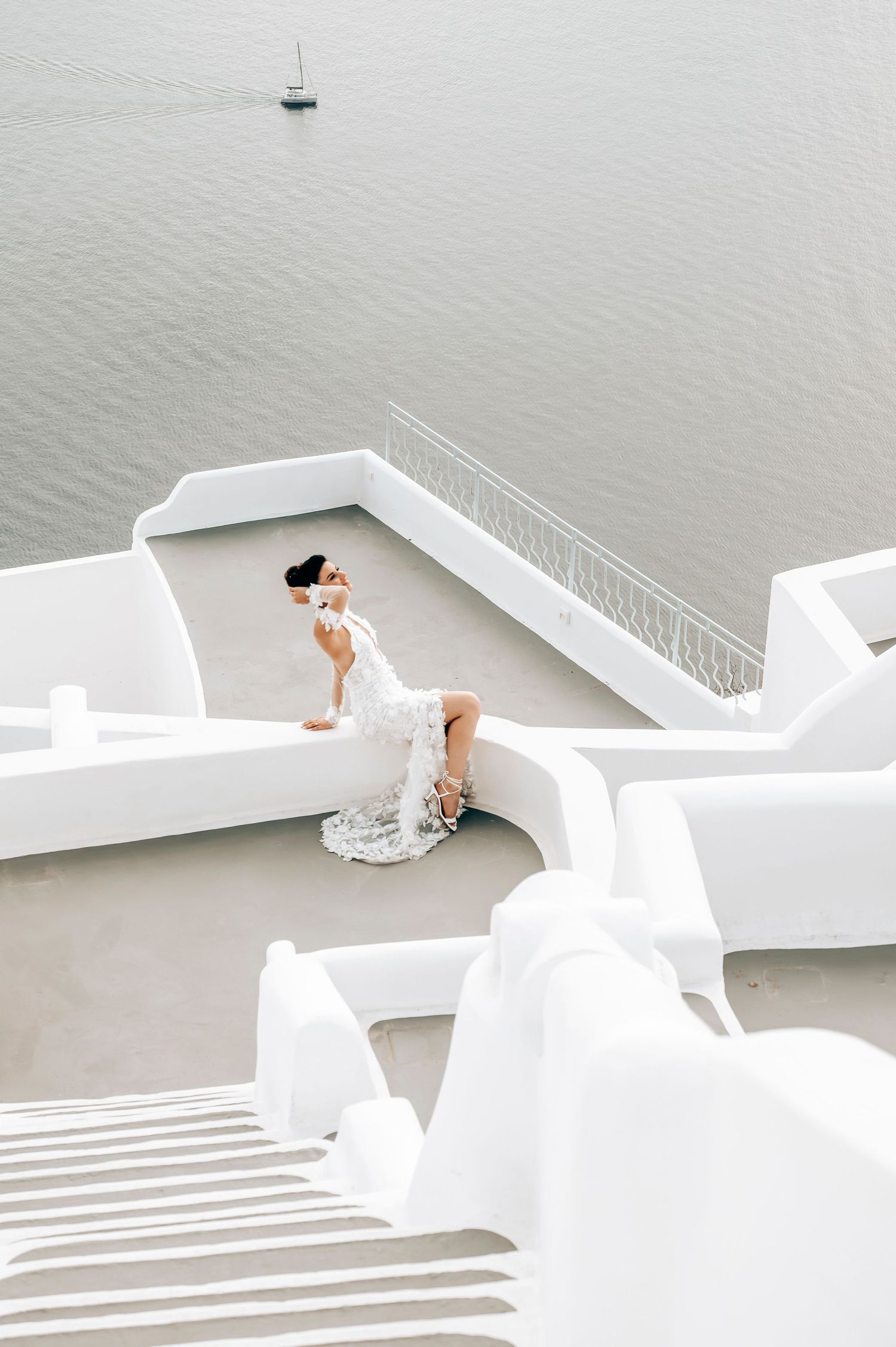 White wedding dress silhouette against Santorini's iconic architecture with sailboat in distance.