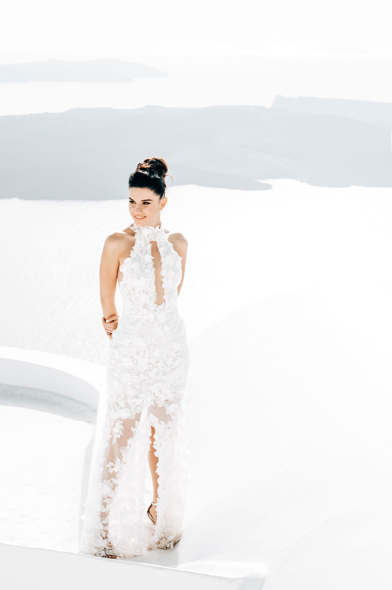 Bride in white lace dress posing on white surface against scenic backdrop.