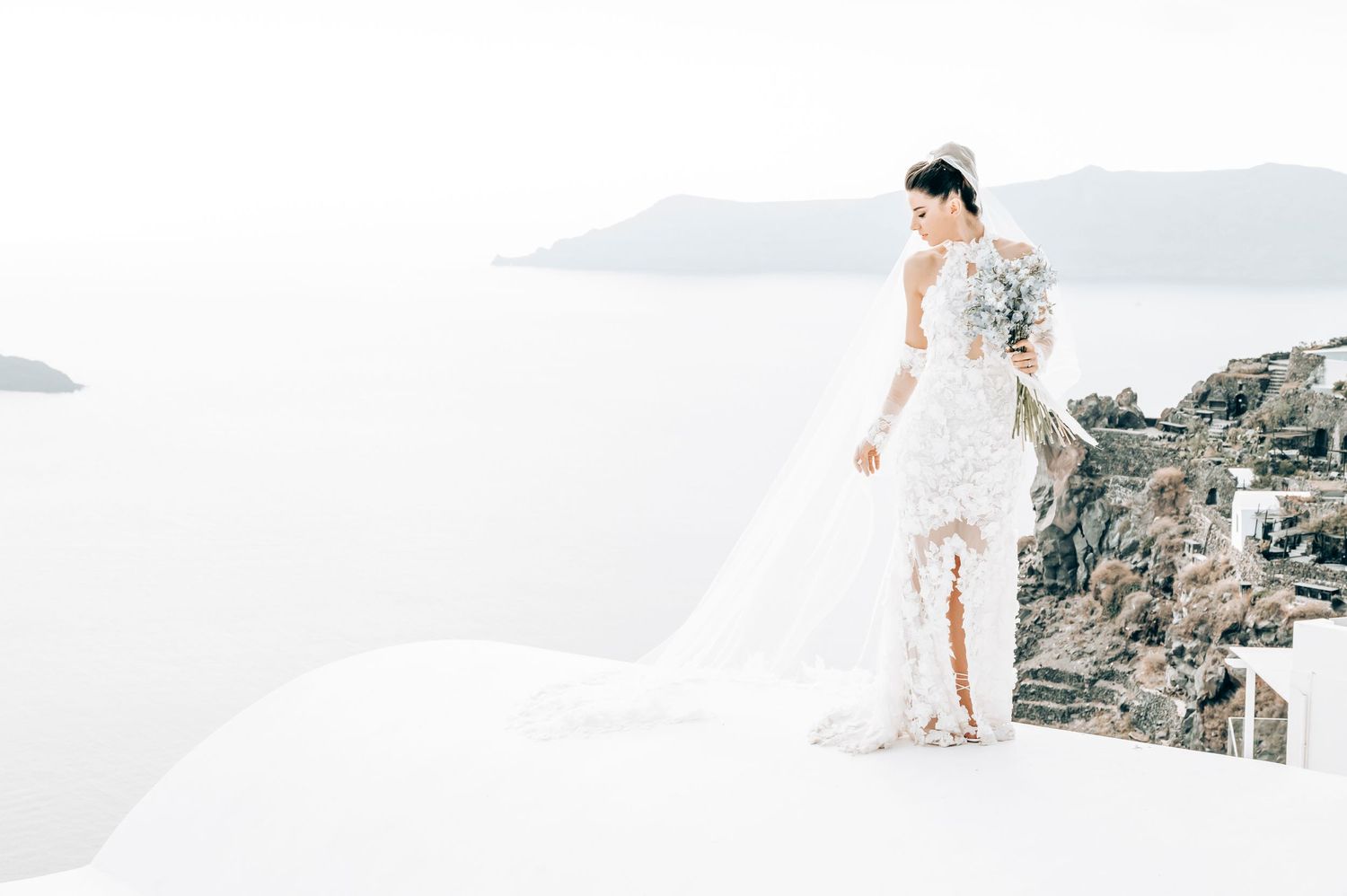 Bride in flowing lace dress overlooking Santorini's white architecture and sea views.