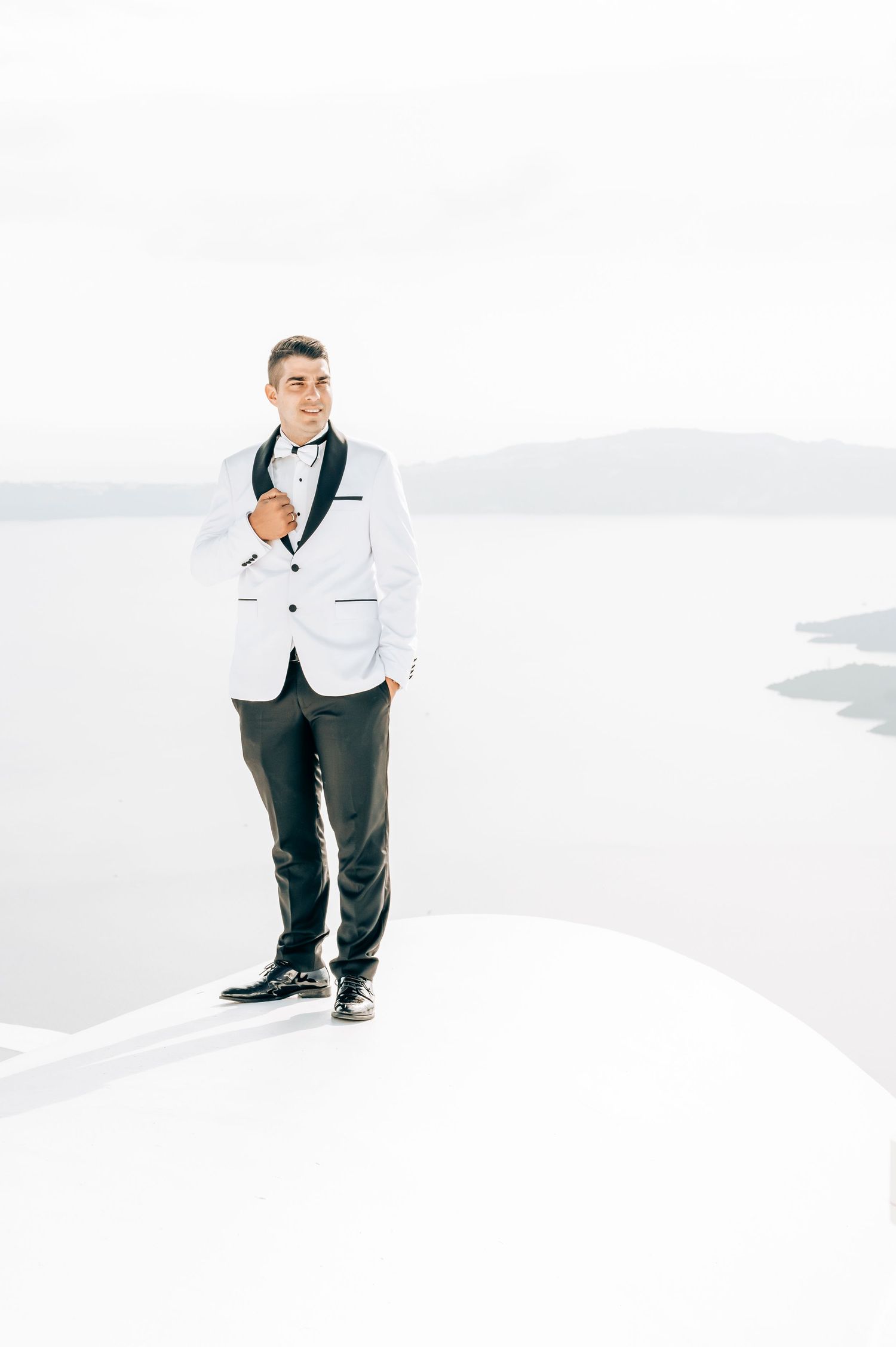 Groom in white tuxedo jacket standing on white surface against scenic background.