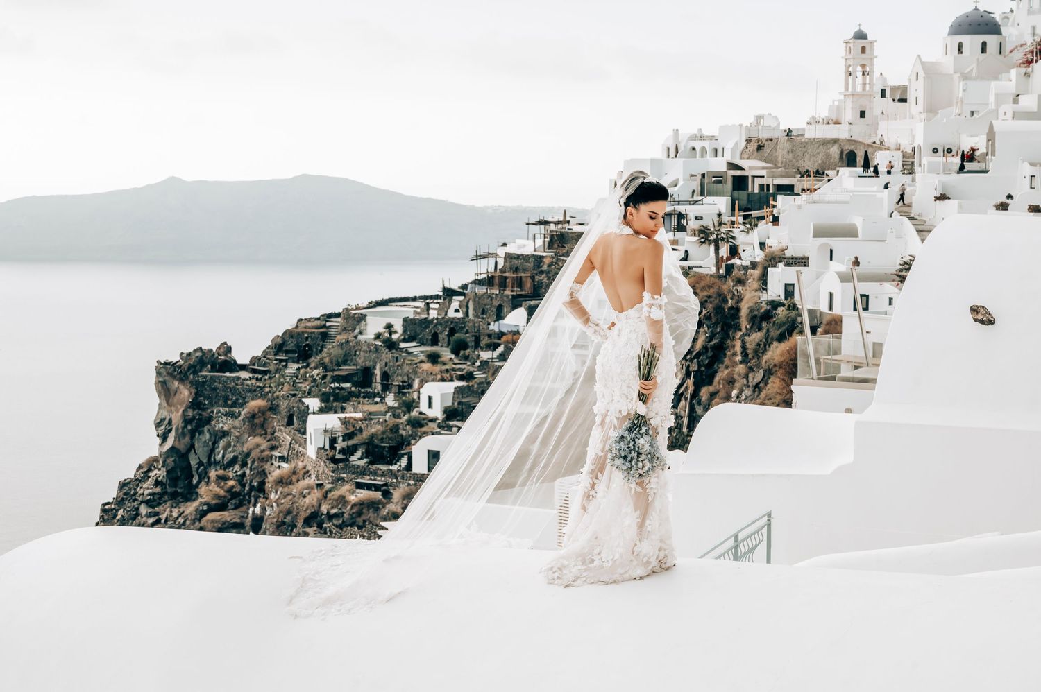 Elegant bridal photo with flowing veil against classic Santorini church and village backdrop.