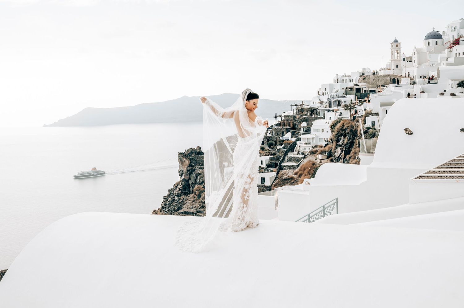 Bride in white lace dress poses on white steps overlooking Santorini's cliffside village.