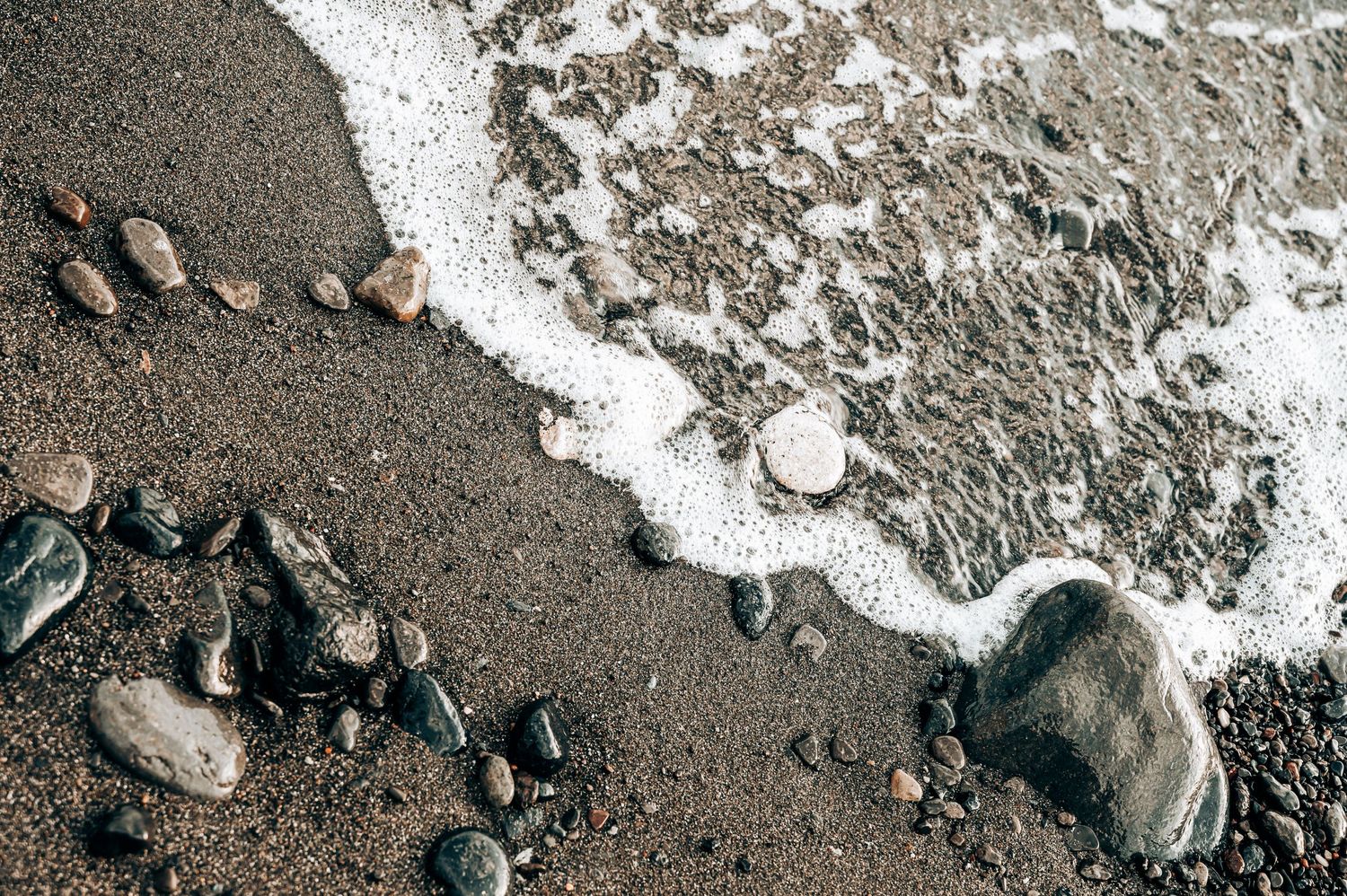Ocean waves washing up on dark volcanic beach with scattered rocks.