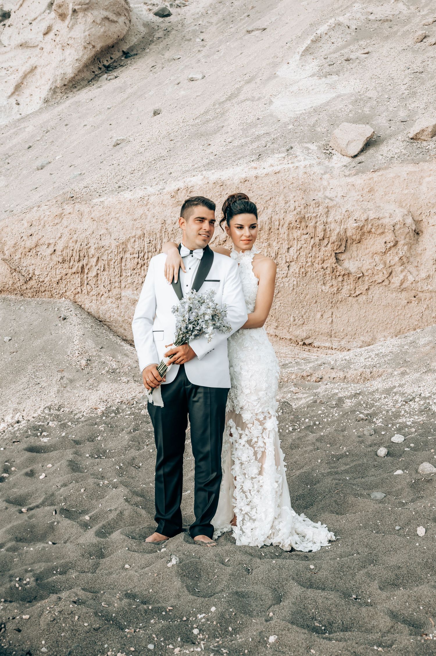 A wedding couple poses on a sandy beach with rocky cliffs in the background.
