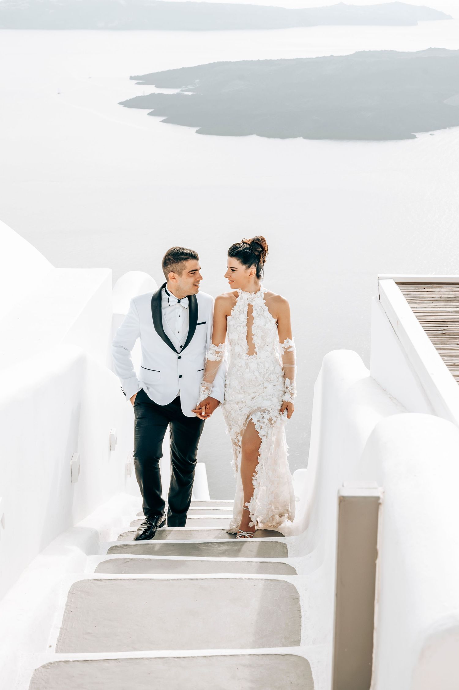 Wedding couple descending white stairs in Santorini with bride in lace dress and groom in white tuxedo jacket.