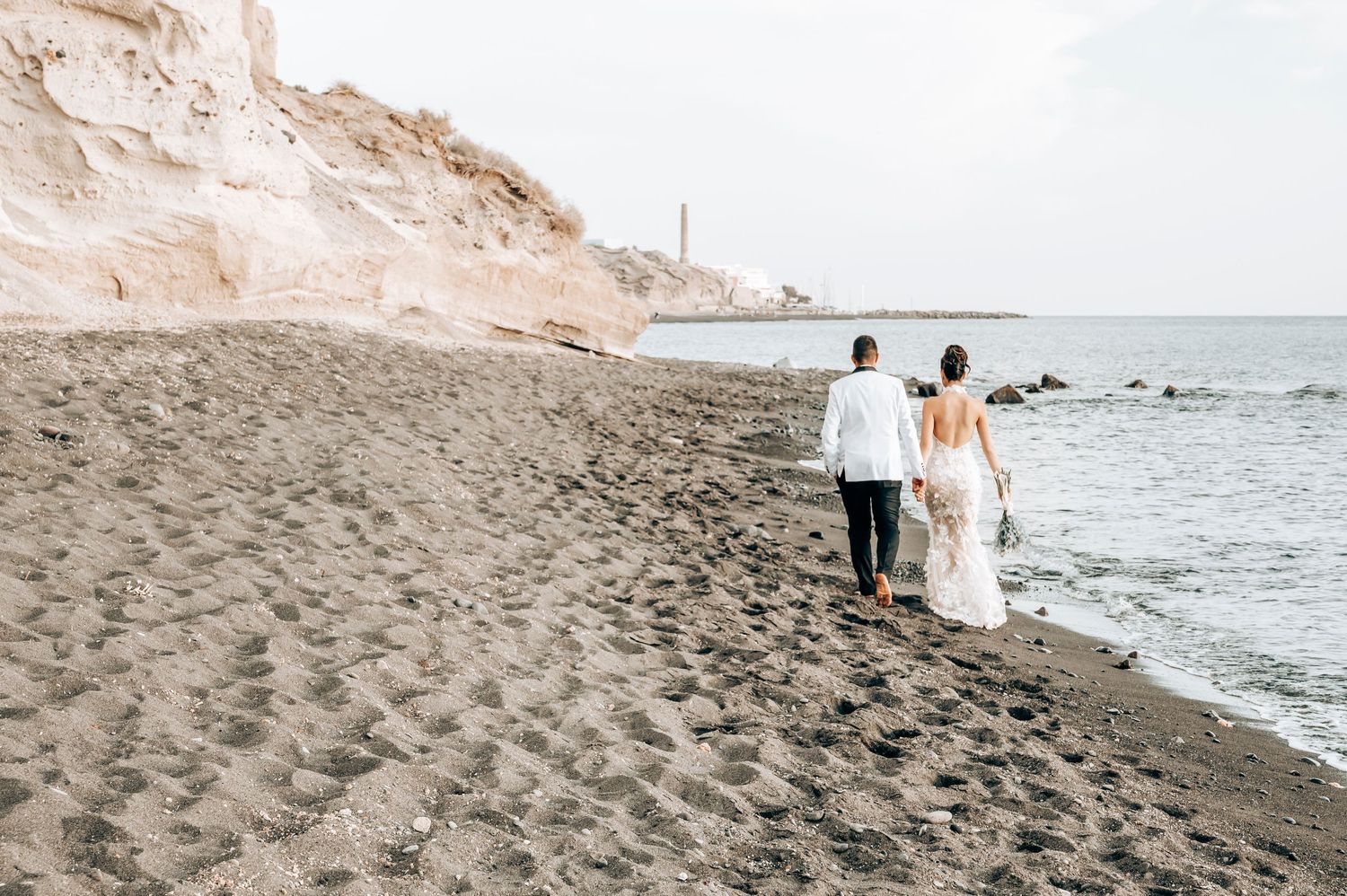 Newlyweds walk hand in hand along a volcanic beach with cliffs in the distance.