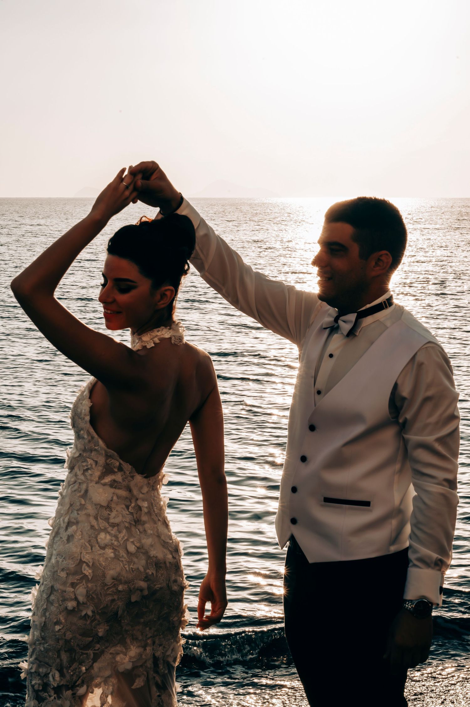 Couple dancing romantically at sunset on a beach with bride in lace dress and groom in white jacket.