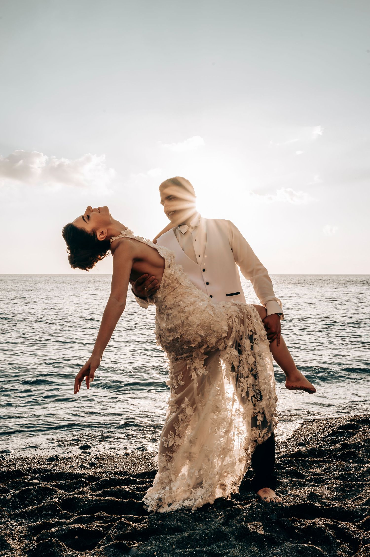 Bride in lace gown dips backwards gracefully against ocean sunset backdrop.