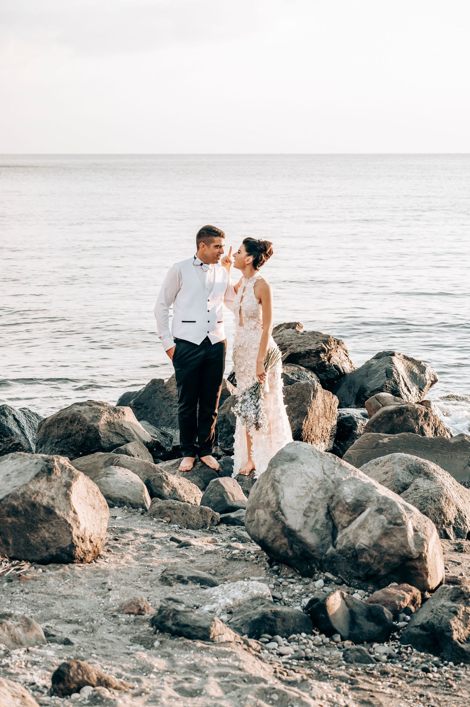 Elegant wedding couple standing on rocky beach overlooking ocean.
