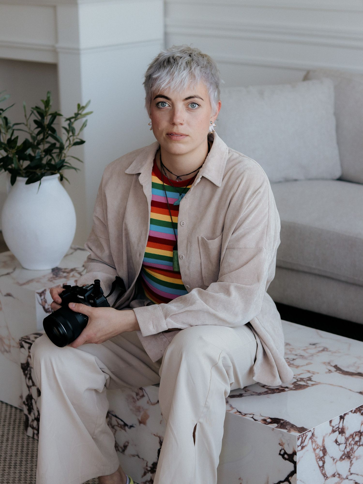 Person in beige outfit sits on white couch in minimalist interior setting with decorative plants.