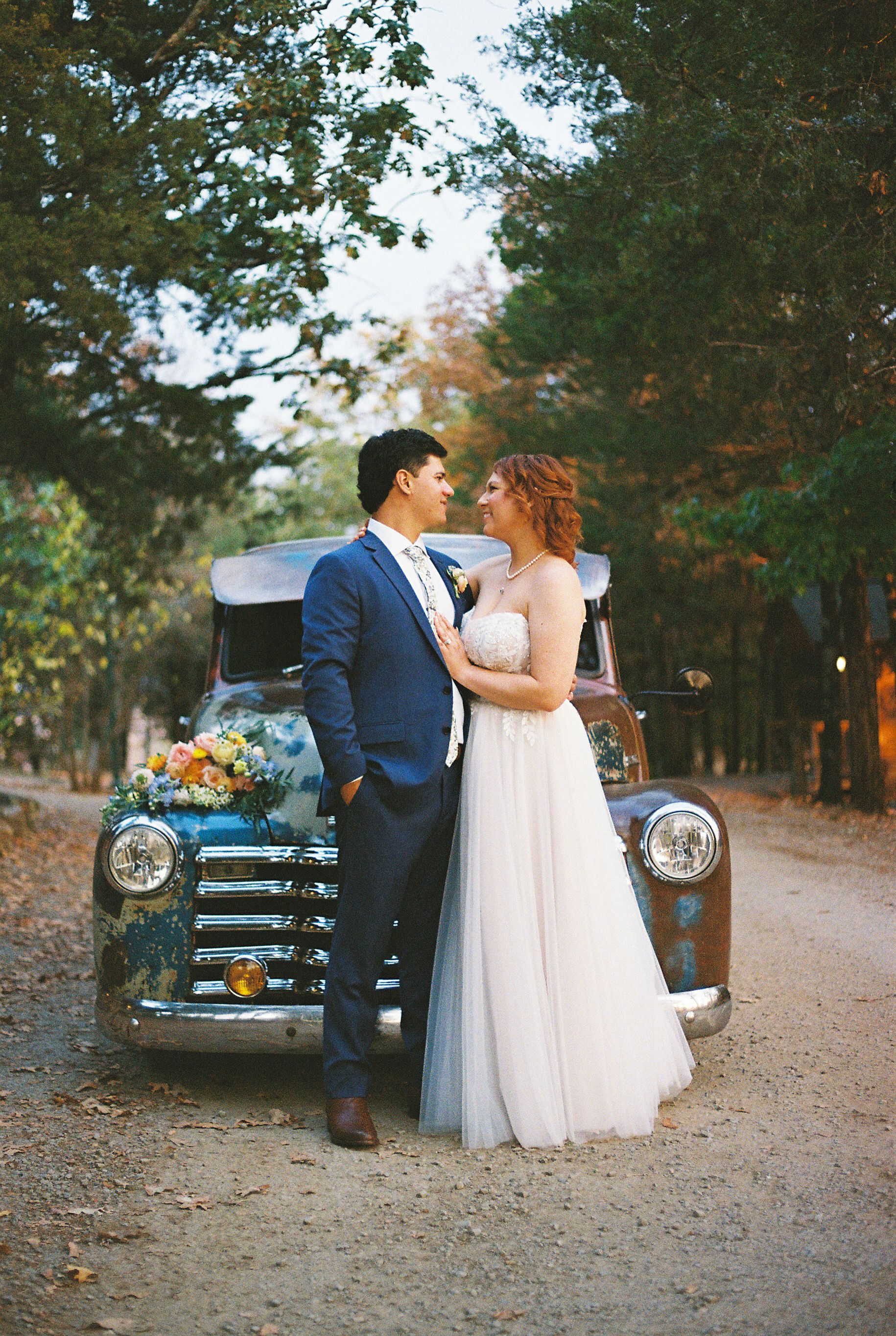 A vintage blue car serves as backdrop for romantic wedding photos on a dirt path.