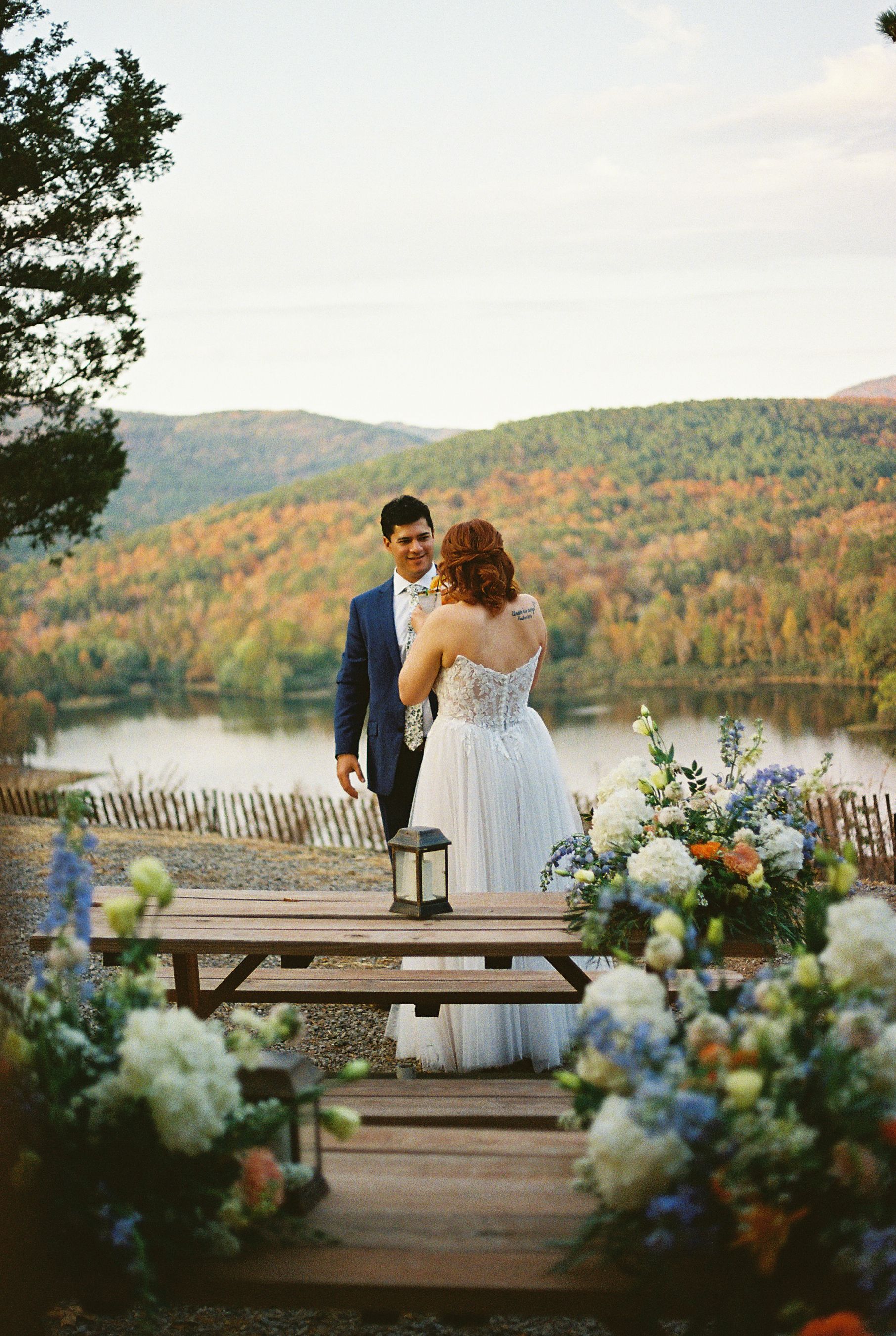 Wedding couple poses on picnic table overlooking autumn lake landscape with floral decorations.