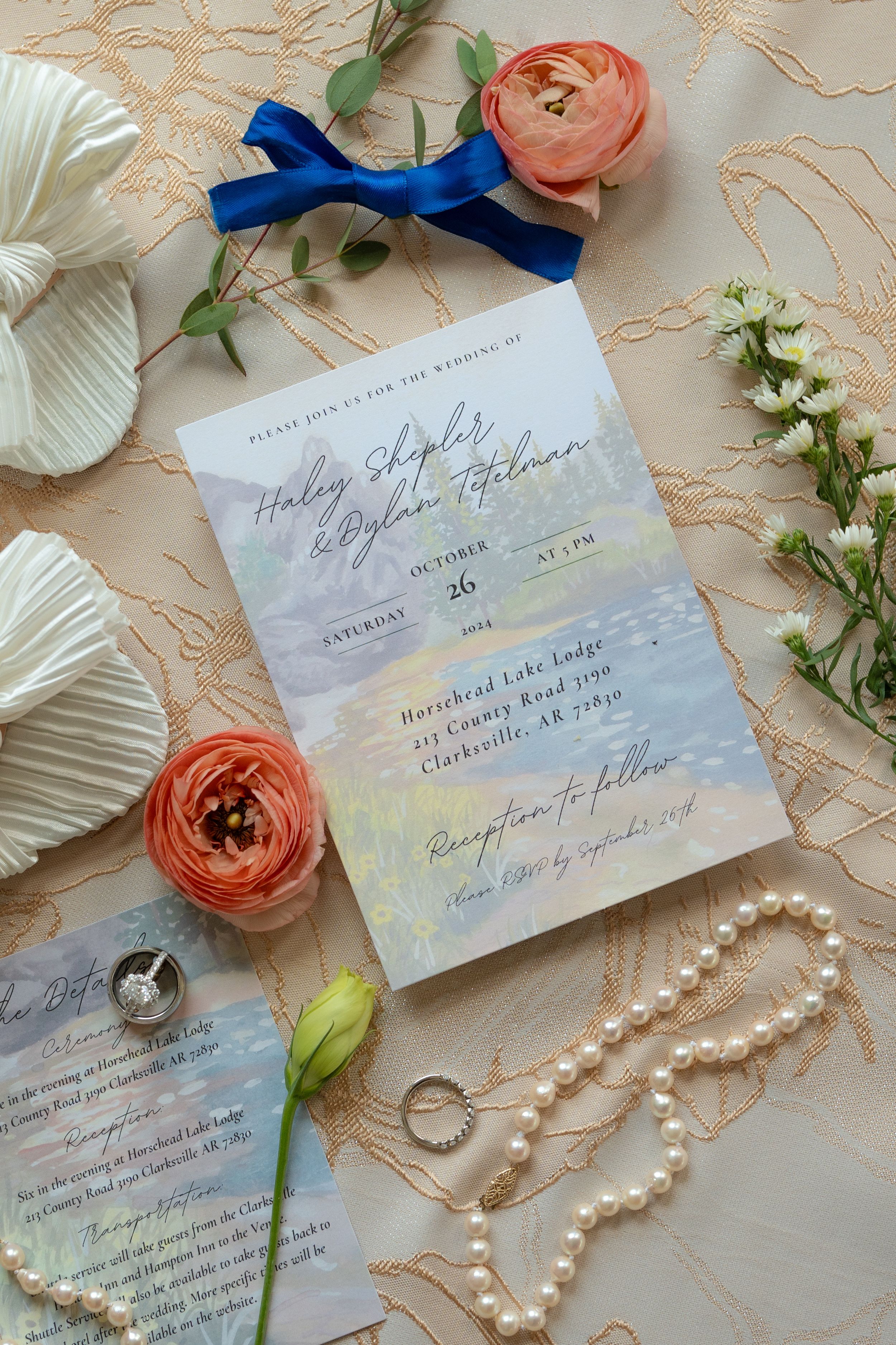 Wedding stationery with pearls, rings, and coral ranunculus flowers arranged on lace fabric.