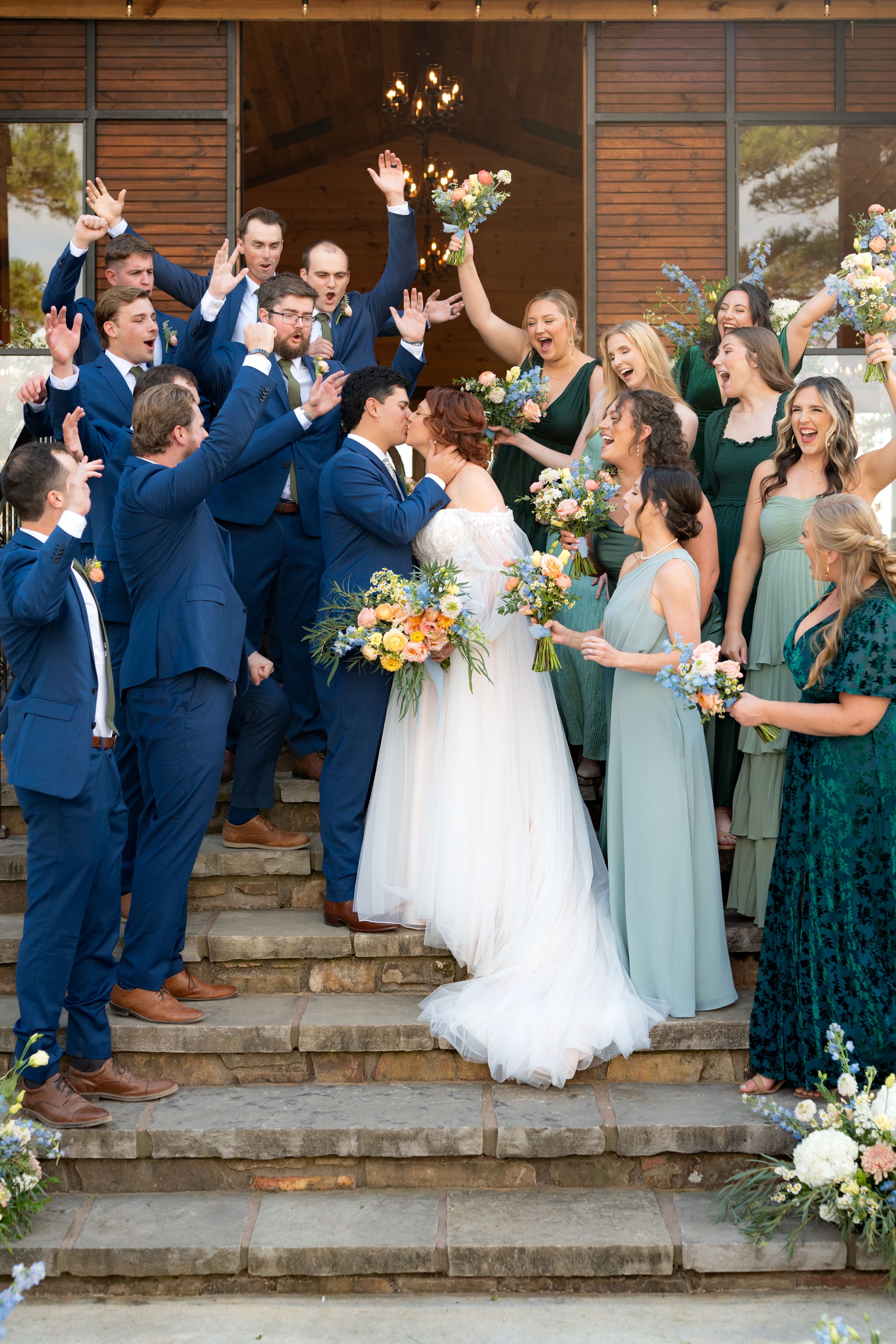 Wedding party celebrates on stone steps with bridesmaids in sage green dresses and groomsmen in navy suits.