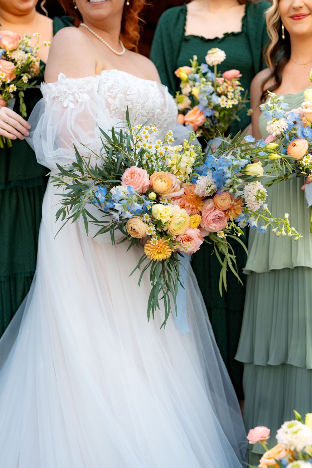 Colorful wedding bouquets featuring peach, blue and yellow flowers held by bridesmaids in green dresses.