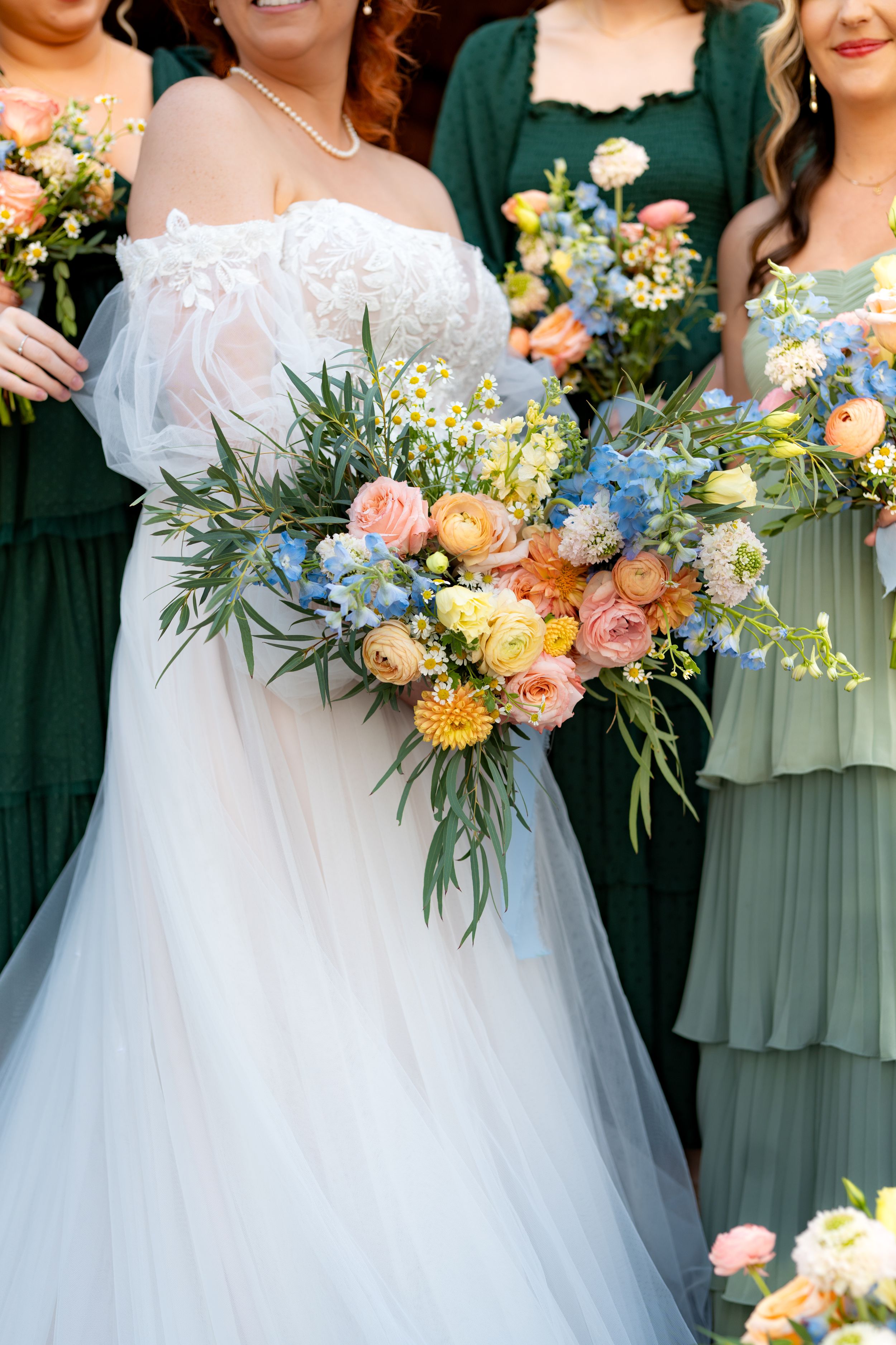 Colorful wedding bouquets featuring peach, blue and yellow flowers held by bridesmaids in green dresses.