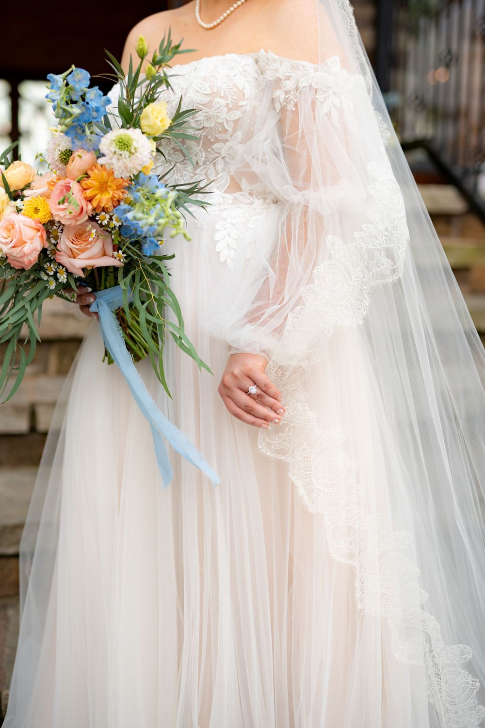A detailed view of a wedding bouquet featuring peach roses, blue delphiniums, and white daisies with flowing ribbons.