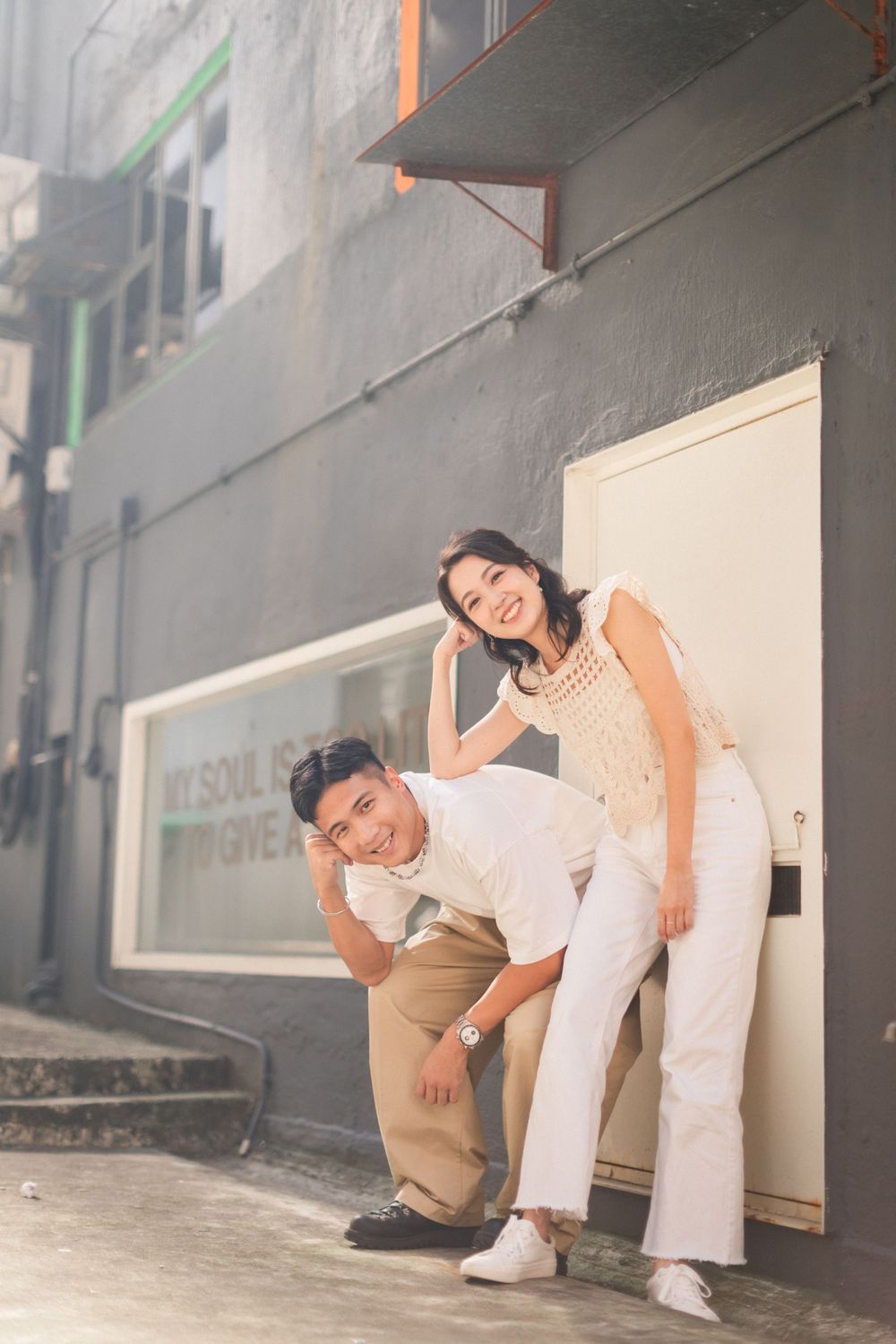 A smiling couple poses playfully outside a gray building, woman leaning on mans back.