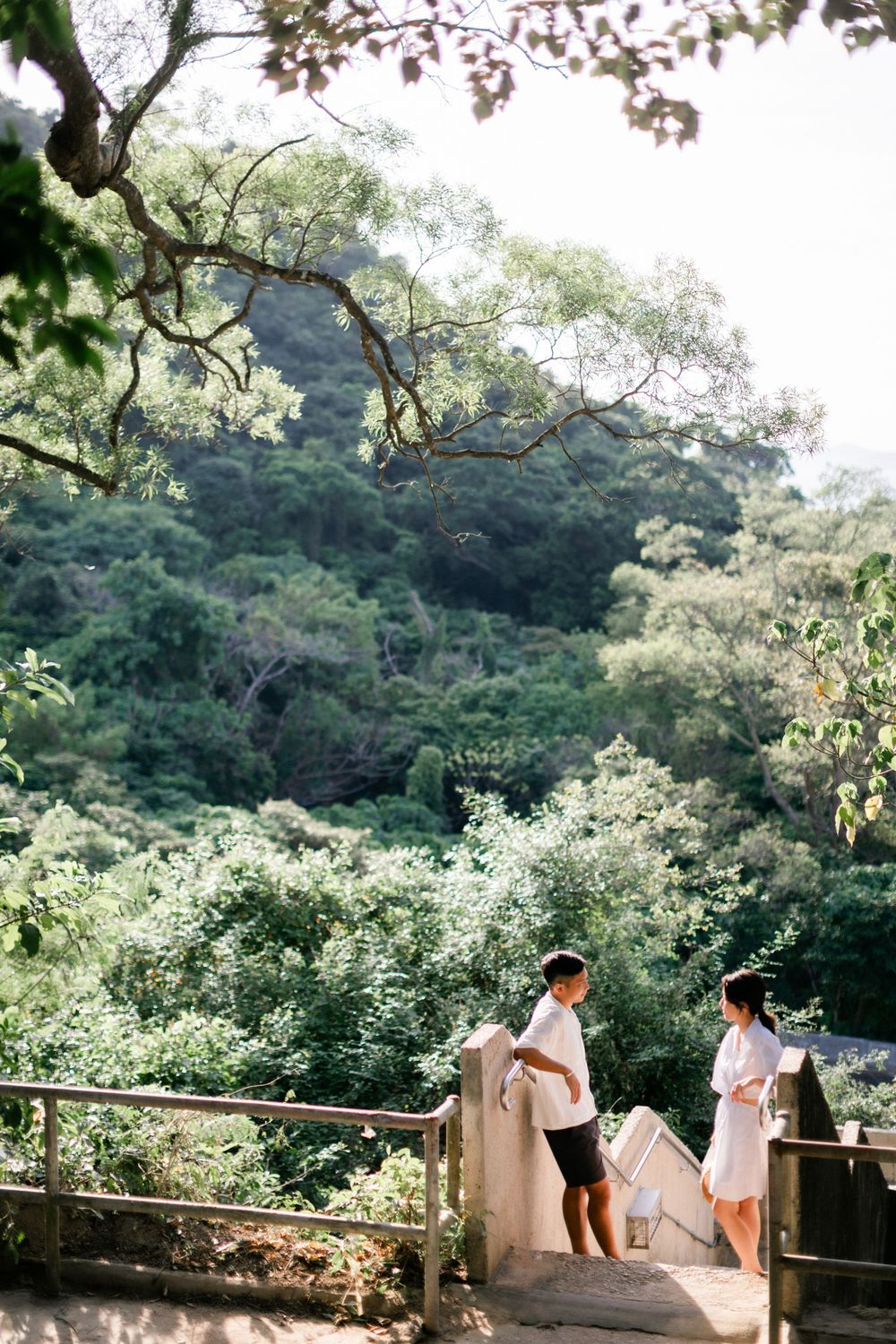 A couple stands on a wooden bridge surrounded by lush green forest and mountains.