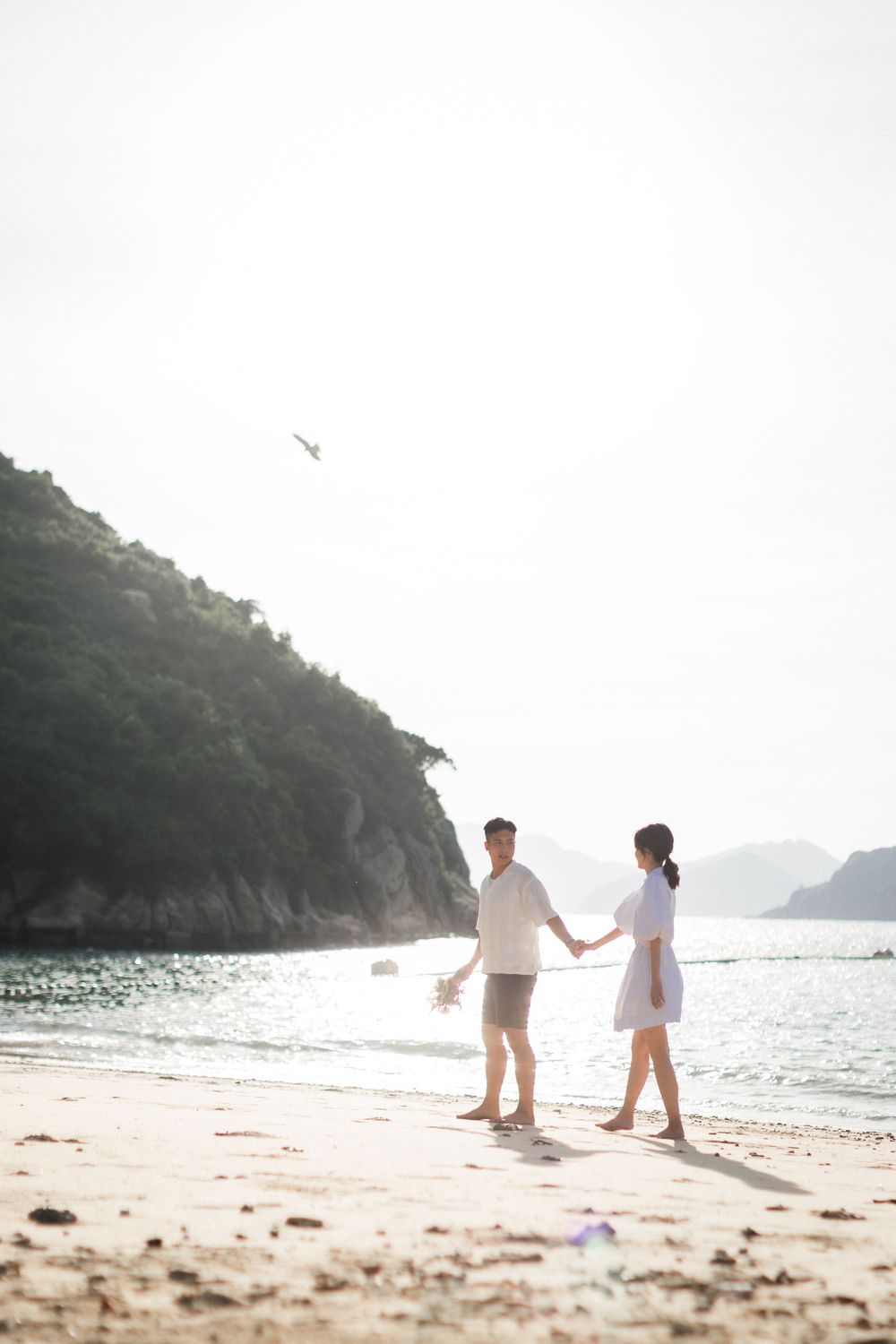 A couple holds hands and walks along a serene beach with mountains in the background.