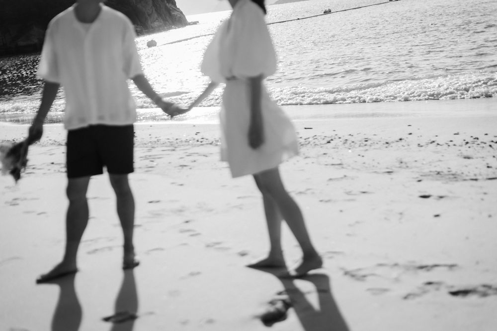 Black and white photo of a couple holding hands while walking barefoot on a beach.
