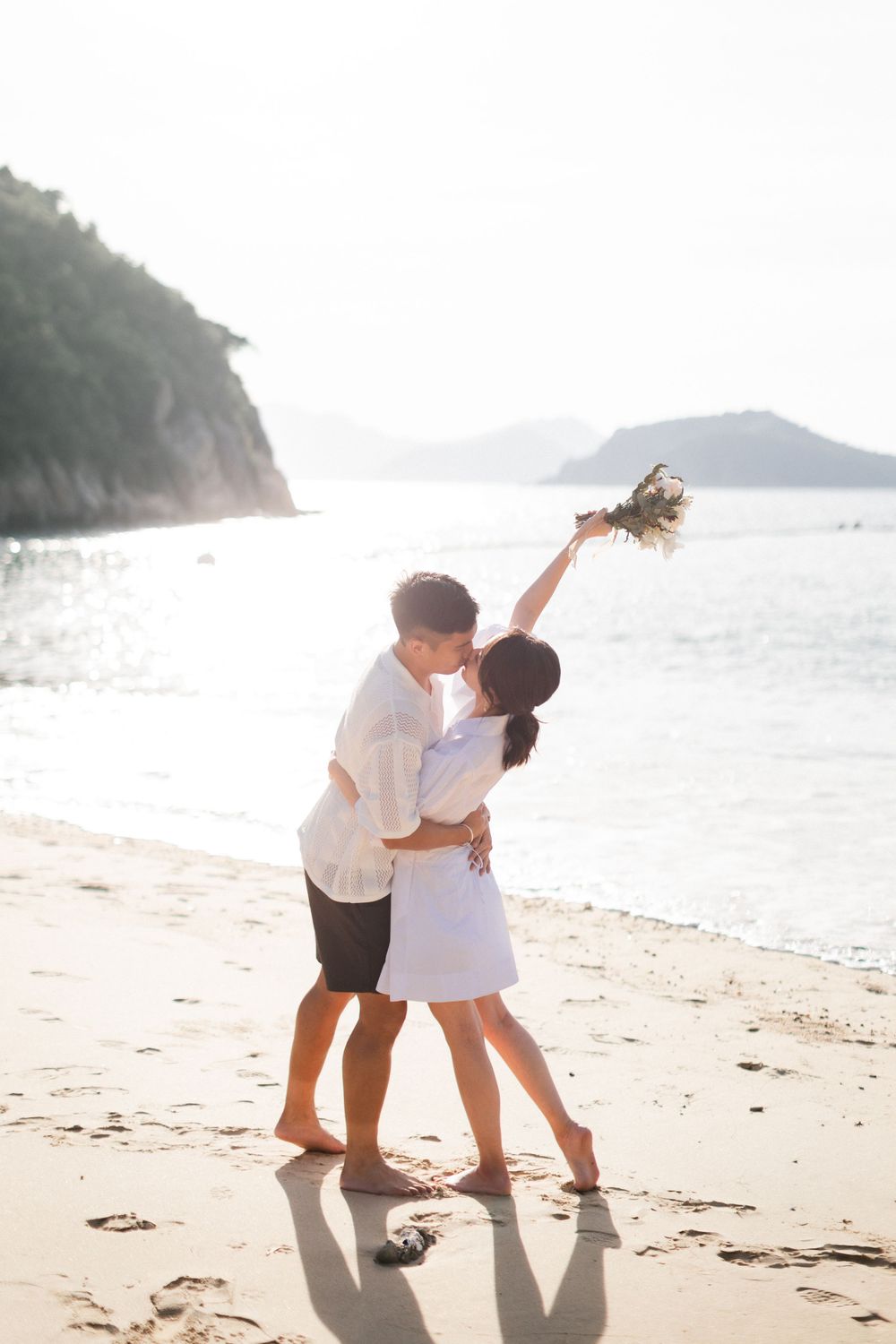 A couple embraces and kisses on a sunny beach, woman raising a floral bouquet joyfully.