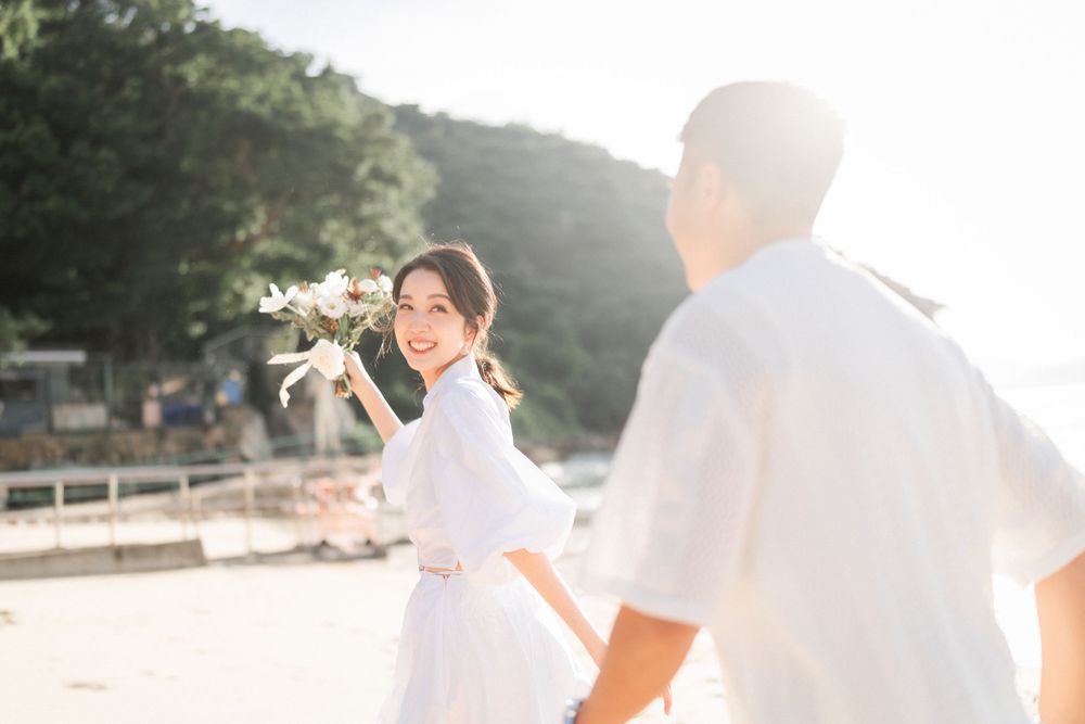 A smiling woman in white holds flowers while holding hands with her partner on a beach.