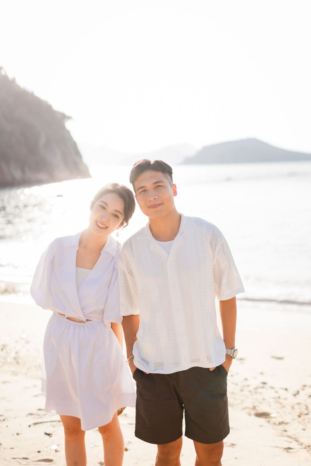 A young Asian couple poses together on a sunny beach, both wearing white outfits with ocean and cliffs behind.