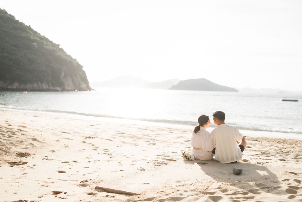 A couple sits together on a sandy beach, gazing at the calm ocean with rocky cliffs in the background.