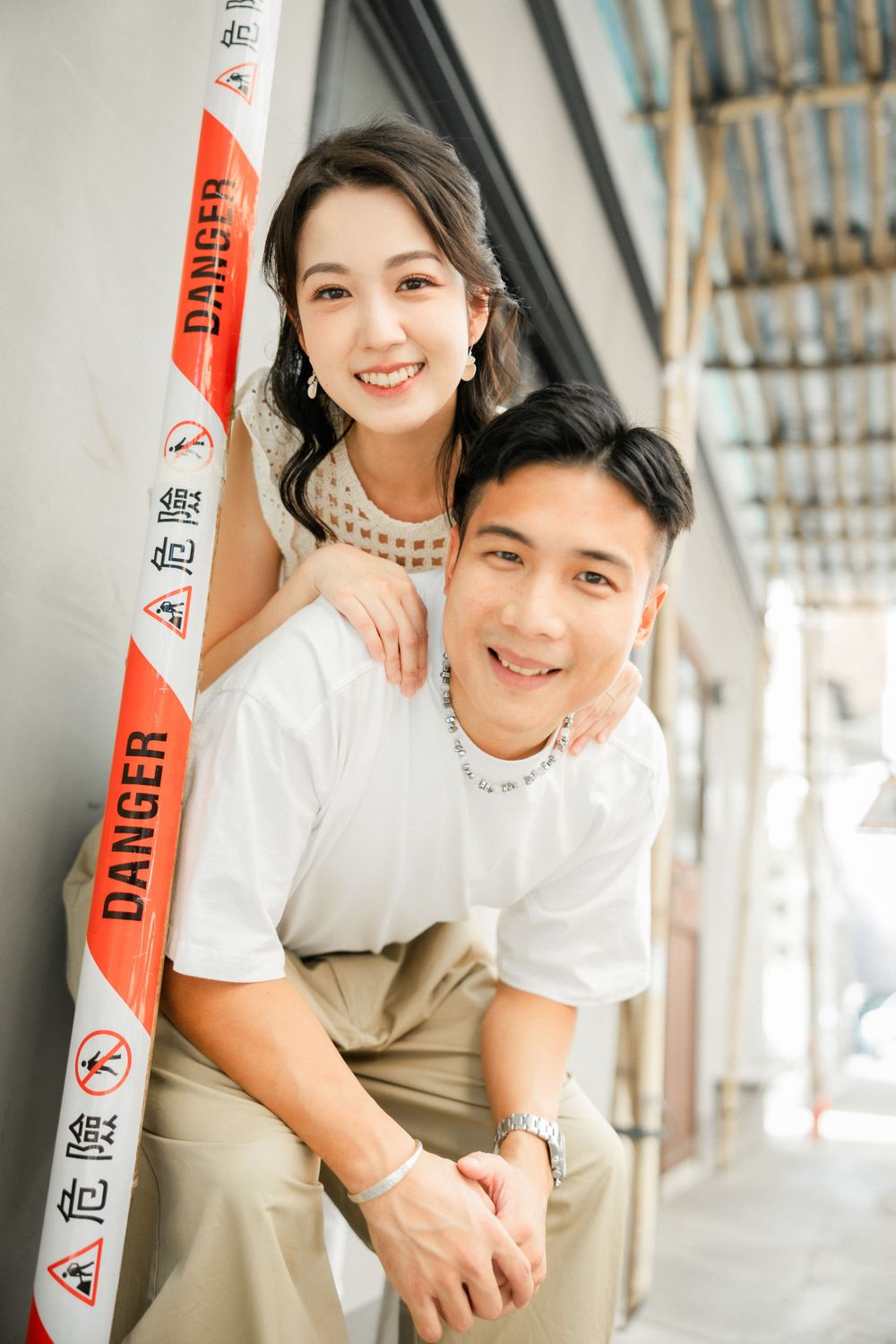 Smiling Asian couple posing together near a danger tape barrier in an urban setting.