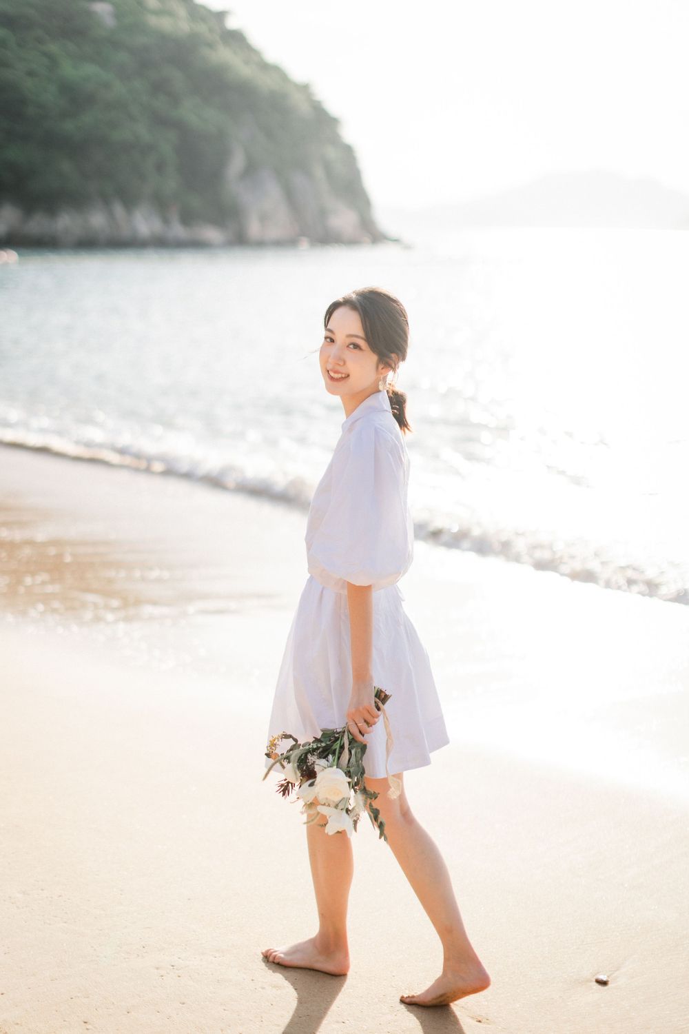A smiling Asian woman in a white dress walks barefoot on a beach, holding a bouquet of white flowers.
