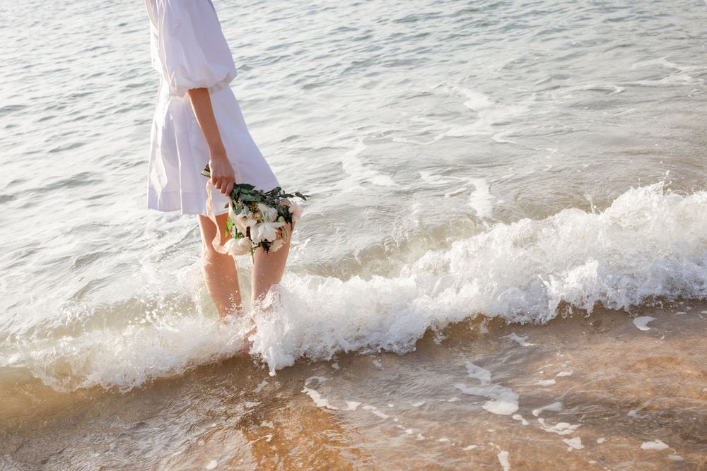 A woman in a white dress stands at the ocean's edge holding a white flower bouquet as waves wash over her feet.