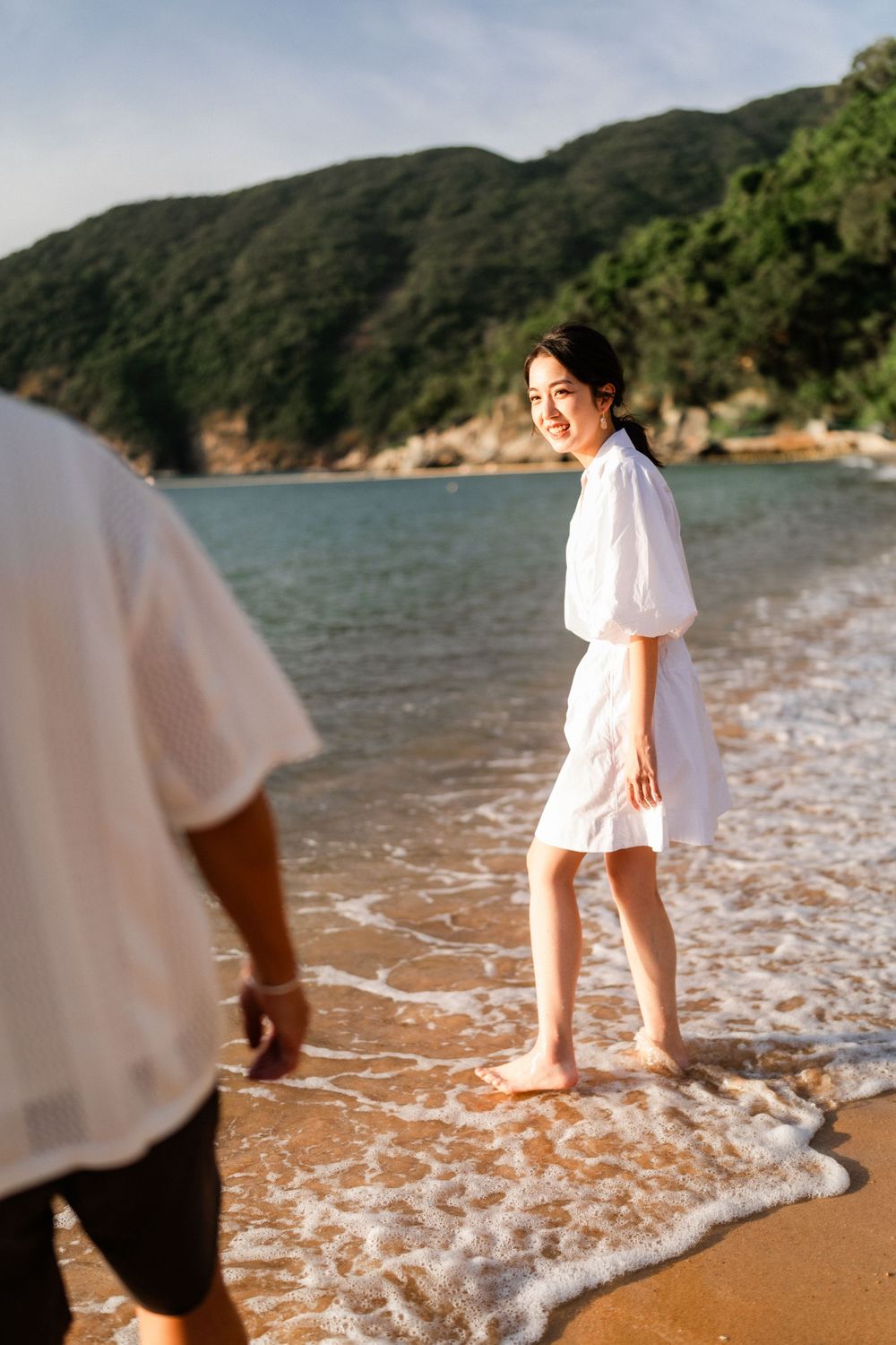 A smiling Asian woman in a white dress stands in ocean waves on a sunny beach with green hills behind her.