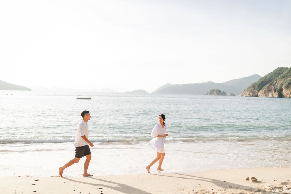 A young Asian couple runs playfully along a sunny beach with blue ocean and green hills in the background.