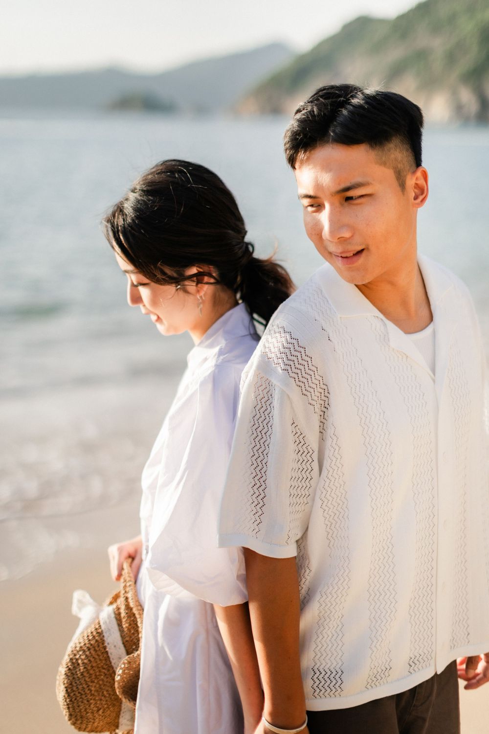 A young Asian couple walks along a sunny beach, the woman carrying a woven bag with ocean waves behind them.