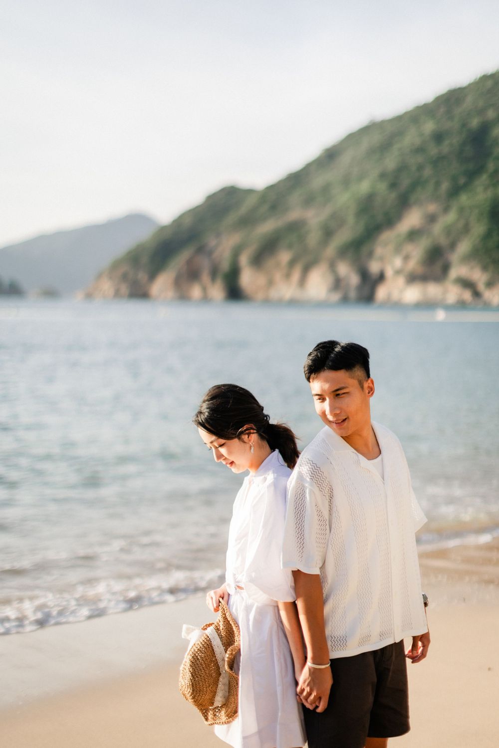 A young Asian couple holds hands while walking on a beach, with blue ocean and green cliffs in the background.