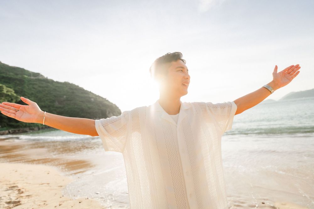 A young Asian man in a white shirt stands on a sunny beach with arms outstretched, enjoying the ocean breeze.
