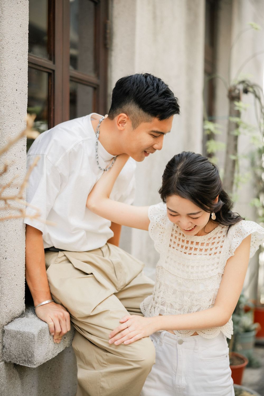 Young Asian couple laughing together outdoors near a rustic building wall with potted plants nearby.
