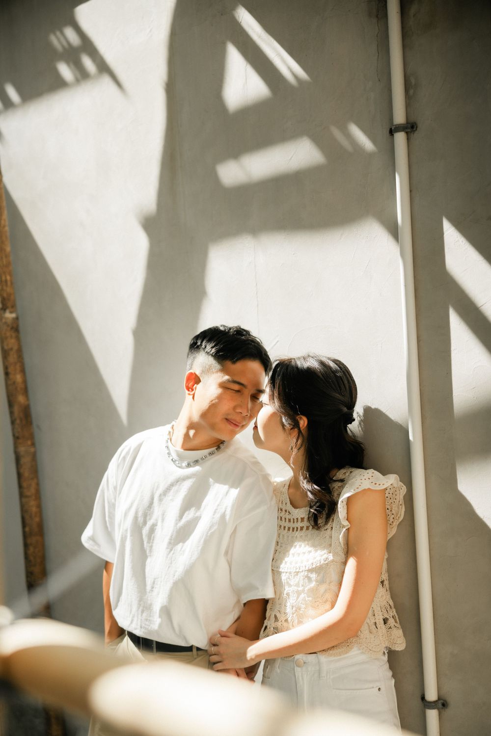 Young Asian couple sharing an intimate moment against a sunlit concrete wall outdoors.