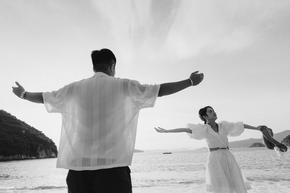 A black and white photo of a couple on a beach with arms outstretched, ocean and hills visible behind them.