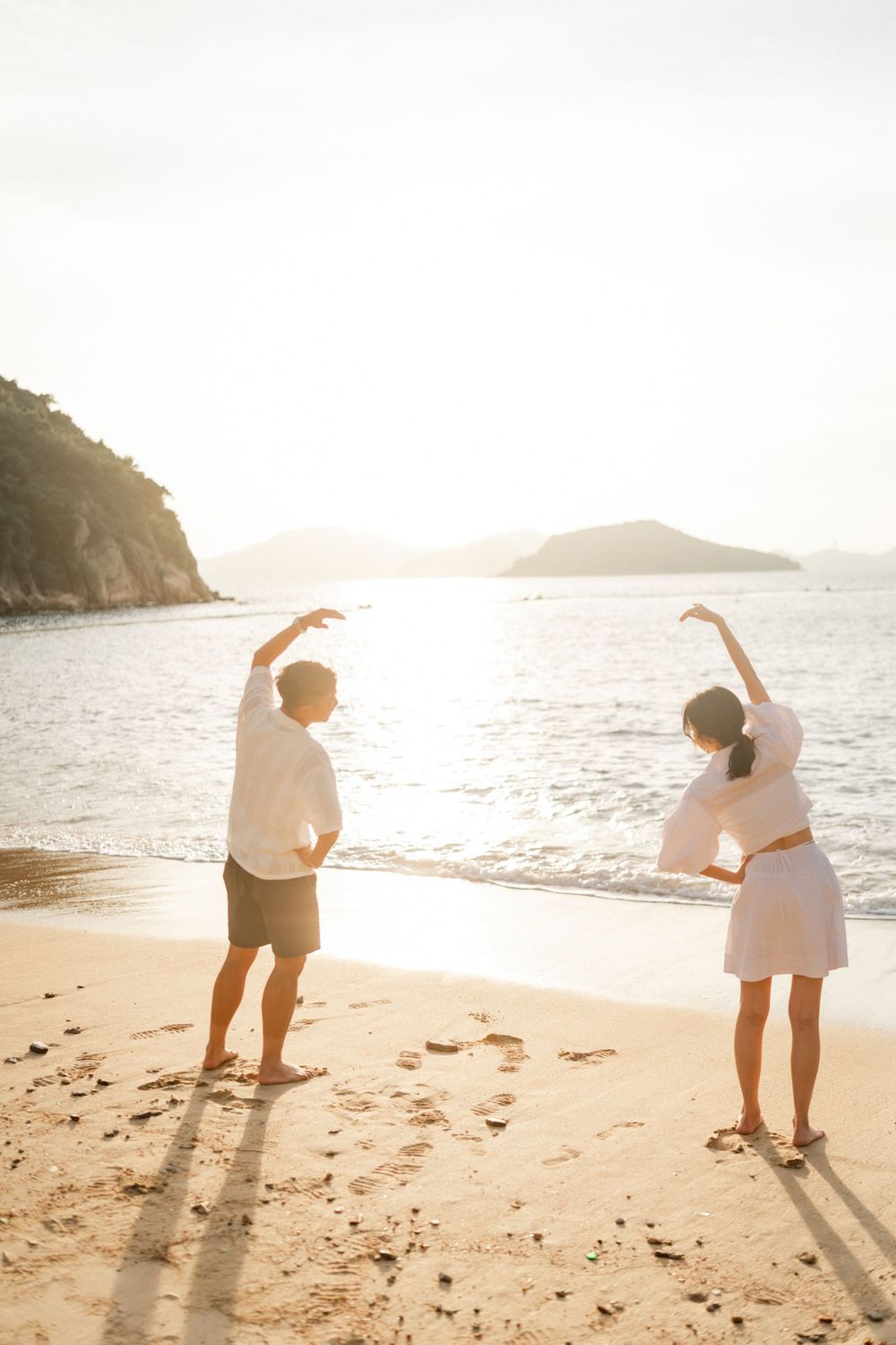 Couple stretching arms up on a golden beach at sunset with scenic ocean and island views.