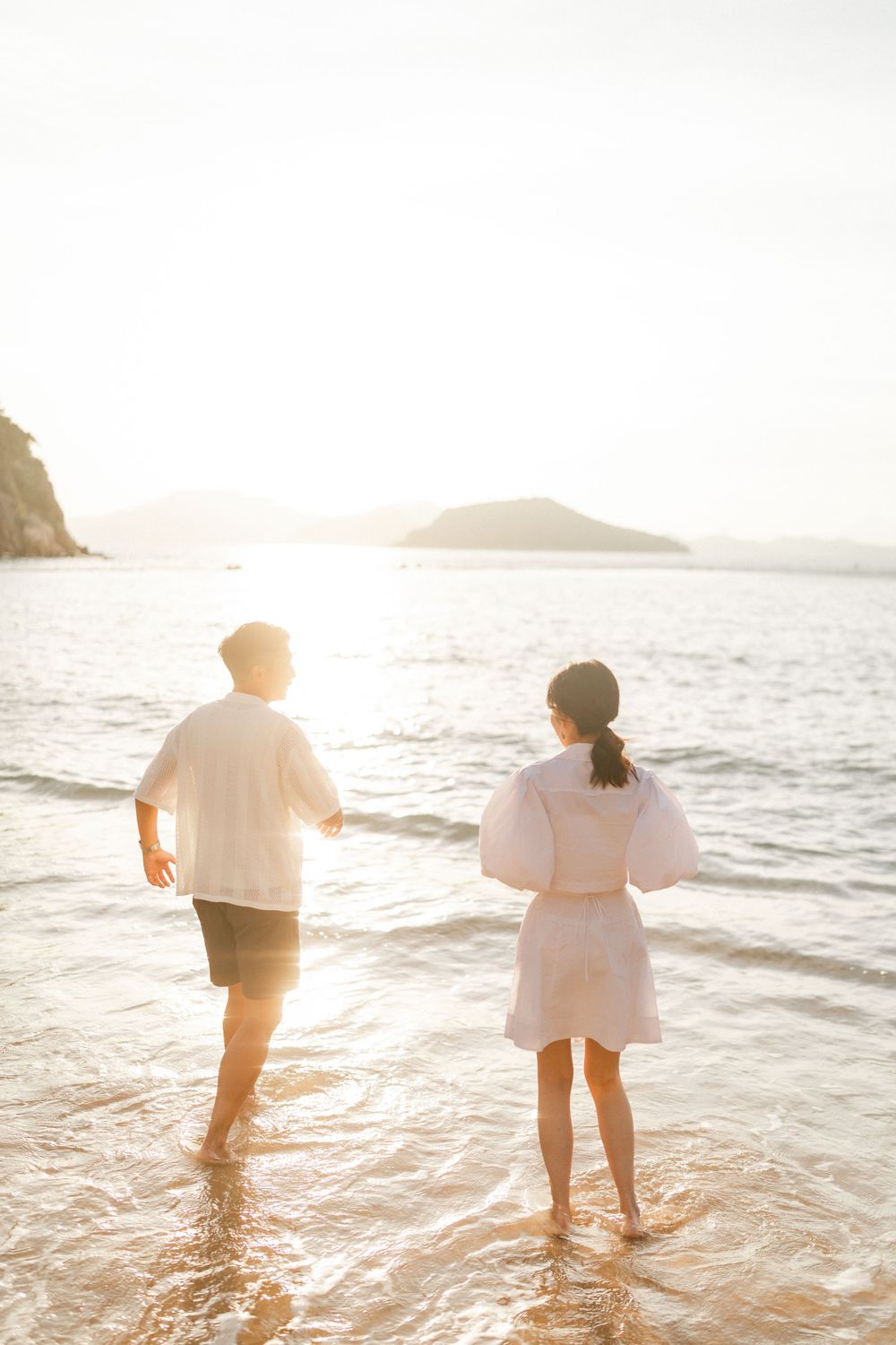Couple holding hands walking into ocean waves at sunset, viewed from behind on a scenic beach.