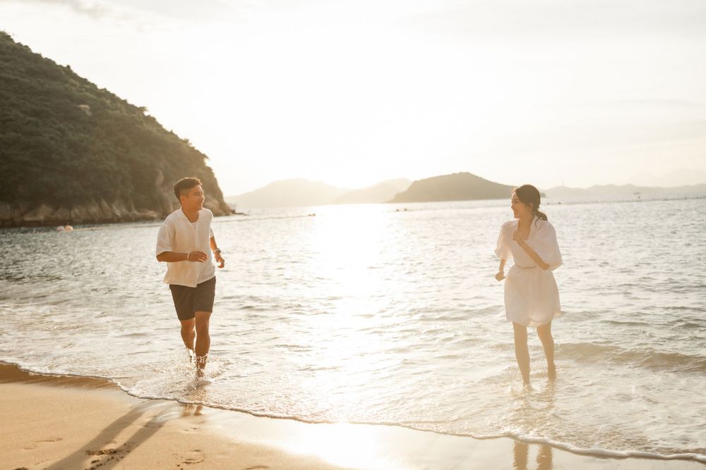 Couple running joyfully along a sandy beach at golden sunset with mountains in background.