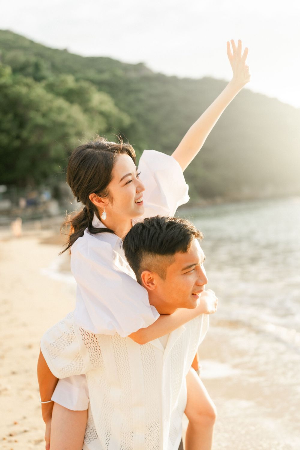 Woman with raised arm enjoying piggyback ride on mans back at sunny beach during sunset.