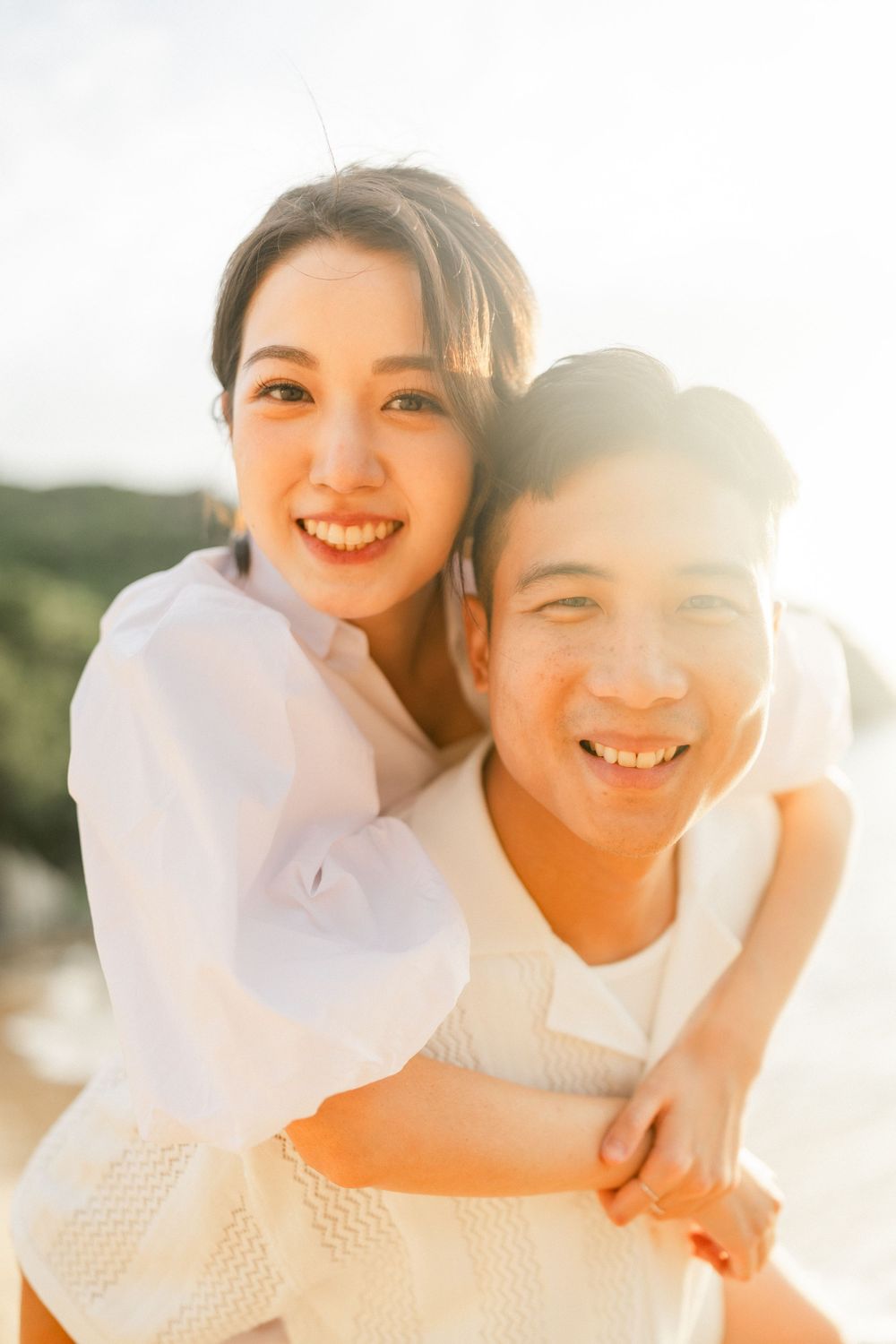Happy Asian couple smiling closely together on beach with warm golden sunset light behind them.