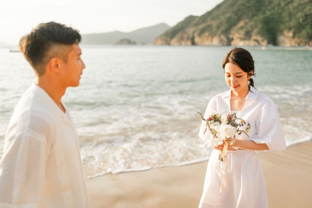 Woman in white dress holding bouquet facing man on sandy beach at golden hour sunset.